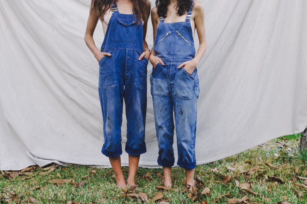 Lifestyle Image by Farhad Samari of two people stand barefoot side by side in overalls in front of a white sheet that is line-drying outdoors